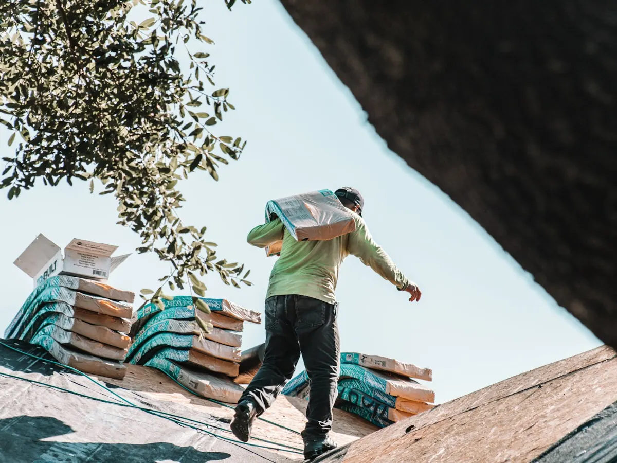 A roofer carrying a bundle of shingles across a roof.
